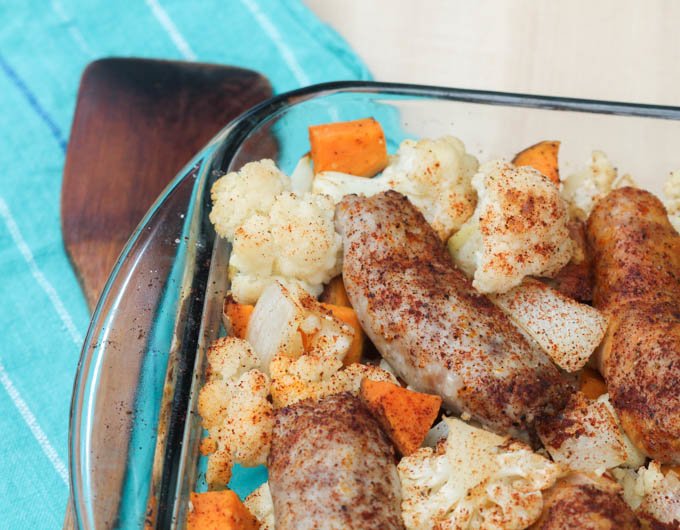 baked sausage, sweet potatoes, and cauliflower in baking dish with wooden spoon and blue placemat