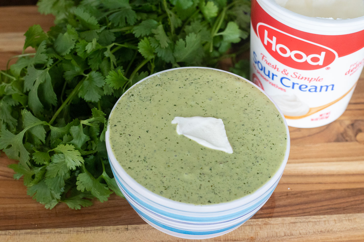 light green creamy sauce with dark green flecks and dollop of sour cream in white and blue striped bowl; bunch of cilantro behind it and white and red container that says Hood sour cream on it.