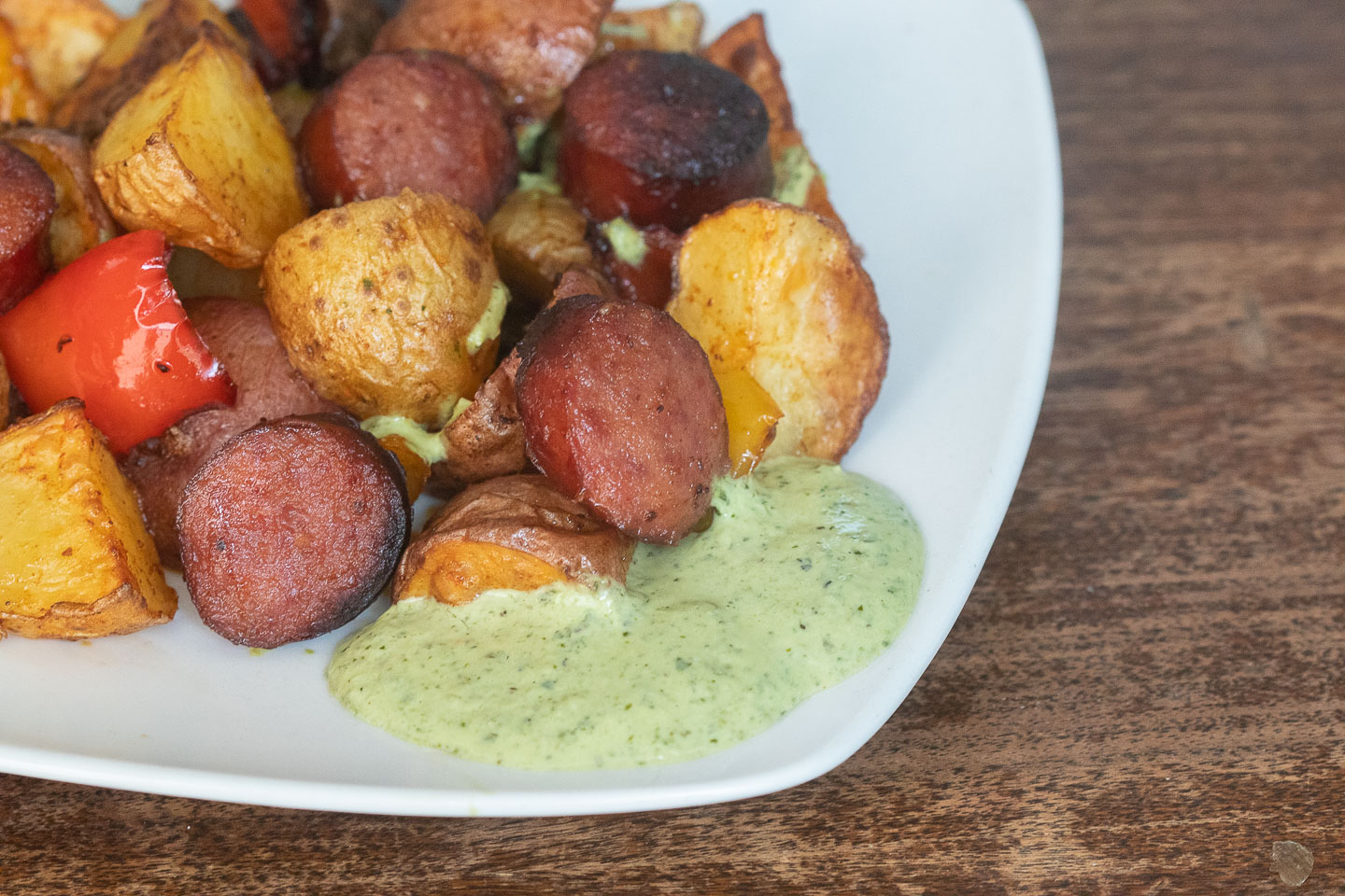baby white potatoes halved and quartered with kielbasa rounds, and red bell pepper on white plate with light green sauce on plate; set on dark wood table